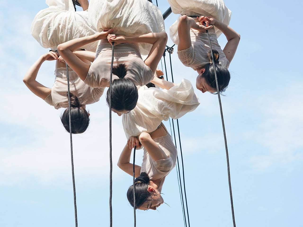 푸른 하늘 아래, 여러 명의 무용수들이 로프에 거꾸로 매달려 공연하고 있다.